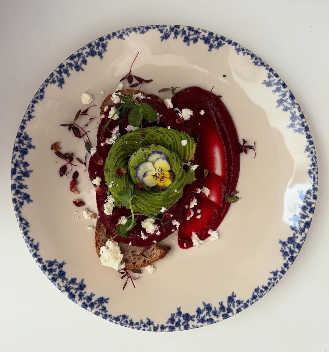 Torrada com creme cor-de-rosa, abacate em forma de flor e queijo feta, num prato de padrão azul e branco, vista de cima