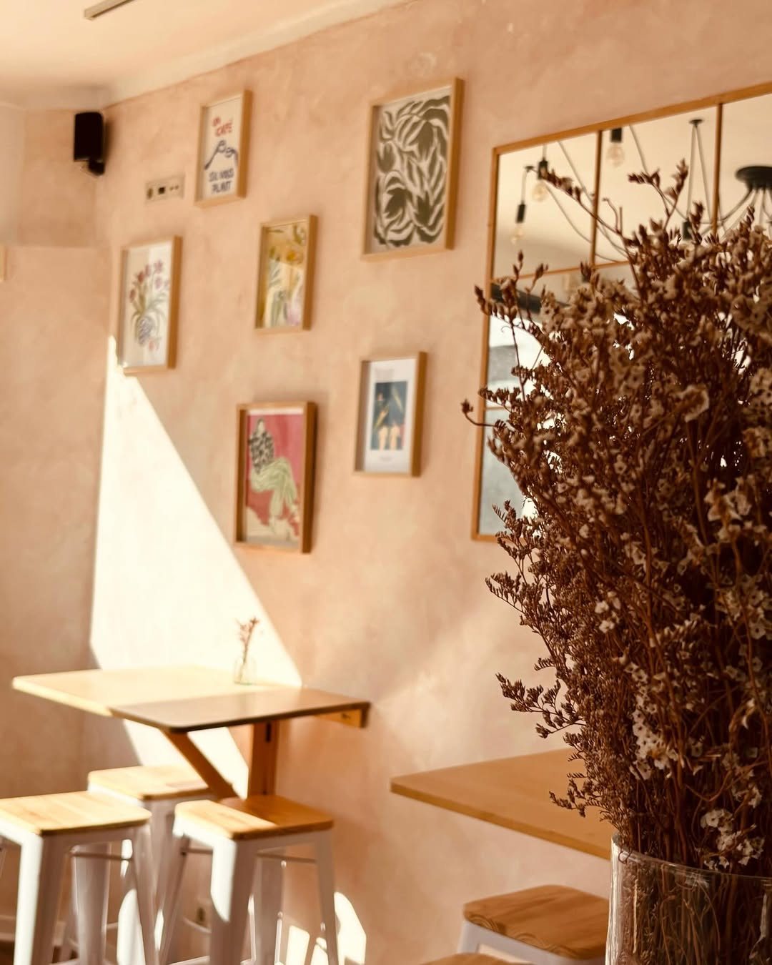 Canto do café com parede cor de salmão, quadros emoldurados, mesas altas de madeira e ramos secos num vaso